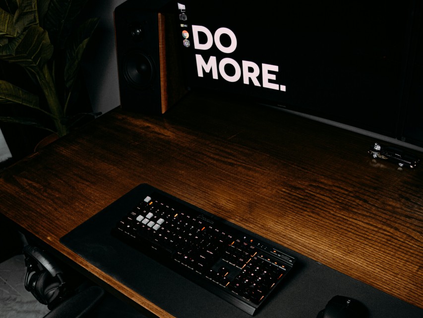 A black computer keyboard on brown wooden desk with the words Do More written on the computer monitor.