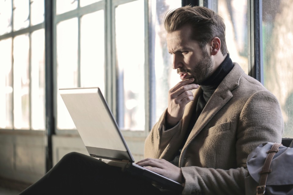 Photo of a man sat by a window in a light coloured suit whilst looking at his laptop confused and stroking his beard