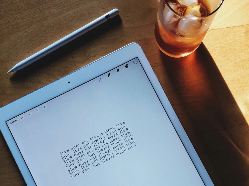 An iPad on a wooden desk with the words ‘Slow does not always mean slow’ written on the screen over and over. With a glass of iced tea alongside the iPad on the desk.