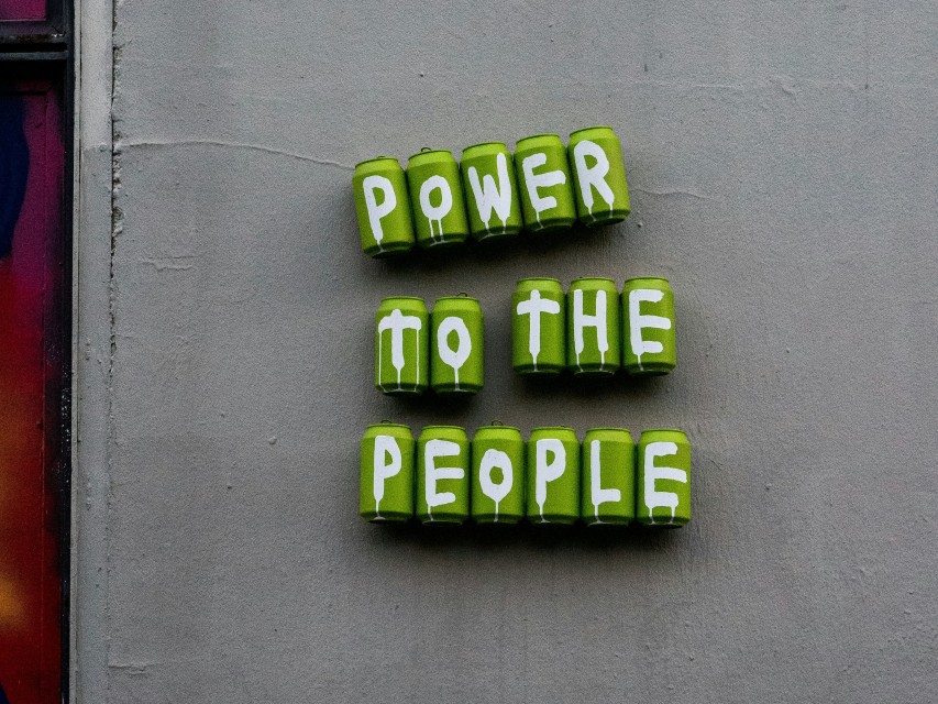 A photo of some drinks cans painted green against a grey wall. The cans have letters spray painted onto them and spell our ‘Power to the people’.