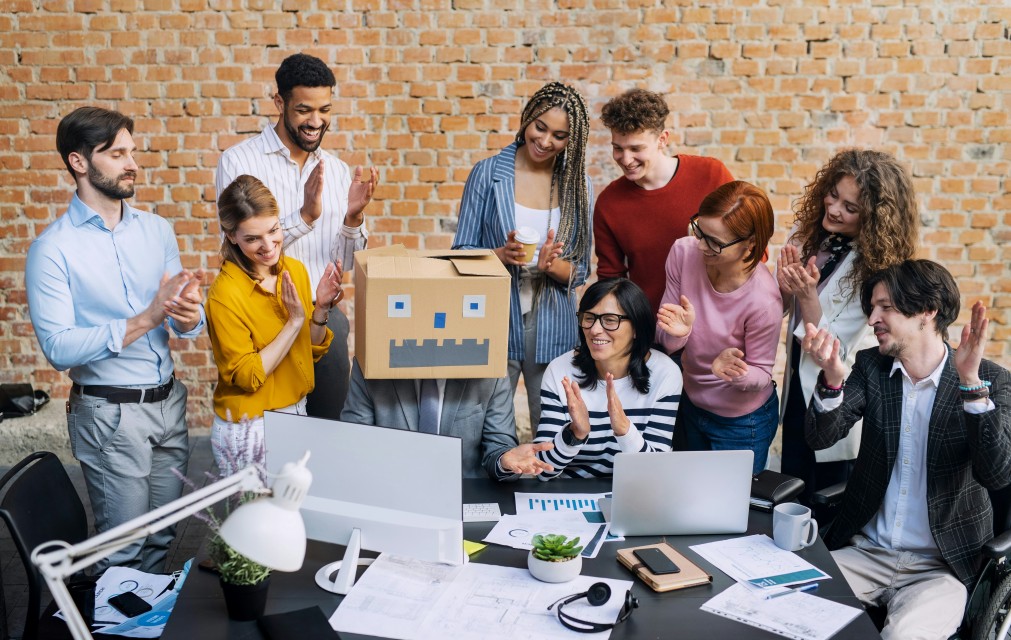 A photo of a group of people having fun together at work, with the person sat at the computer wearing a cardboard robot head.