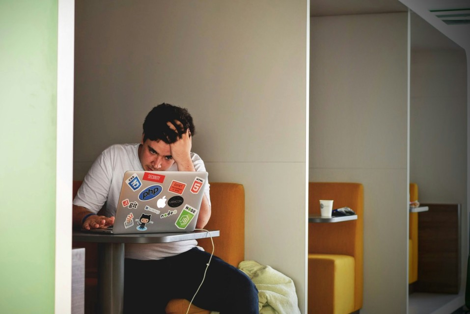 A photo of an engineer working at their laptop looking confused, they are sat in a small booth in an empty office.