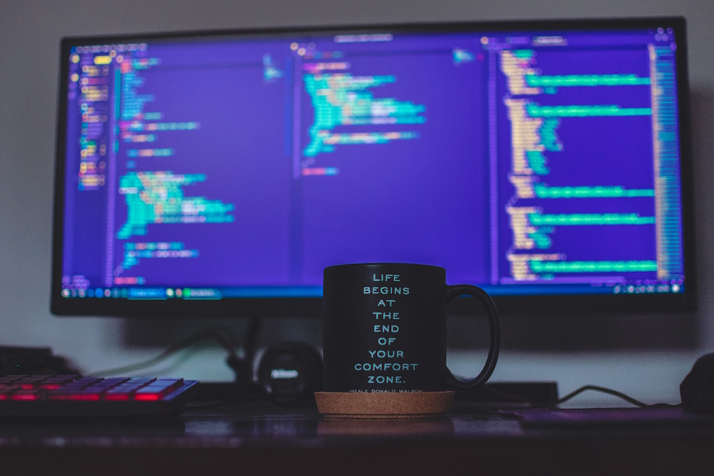 A photo of a coffee cup infront of a coders screens, the cup says Life Begins At The End Of Your Comfort Zone.